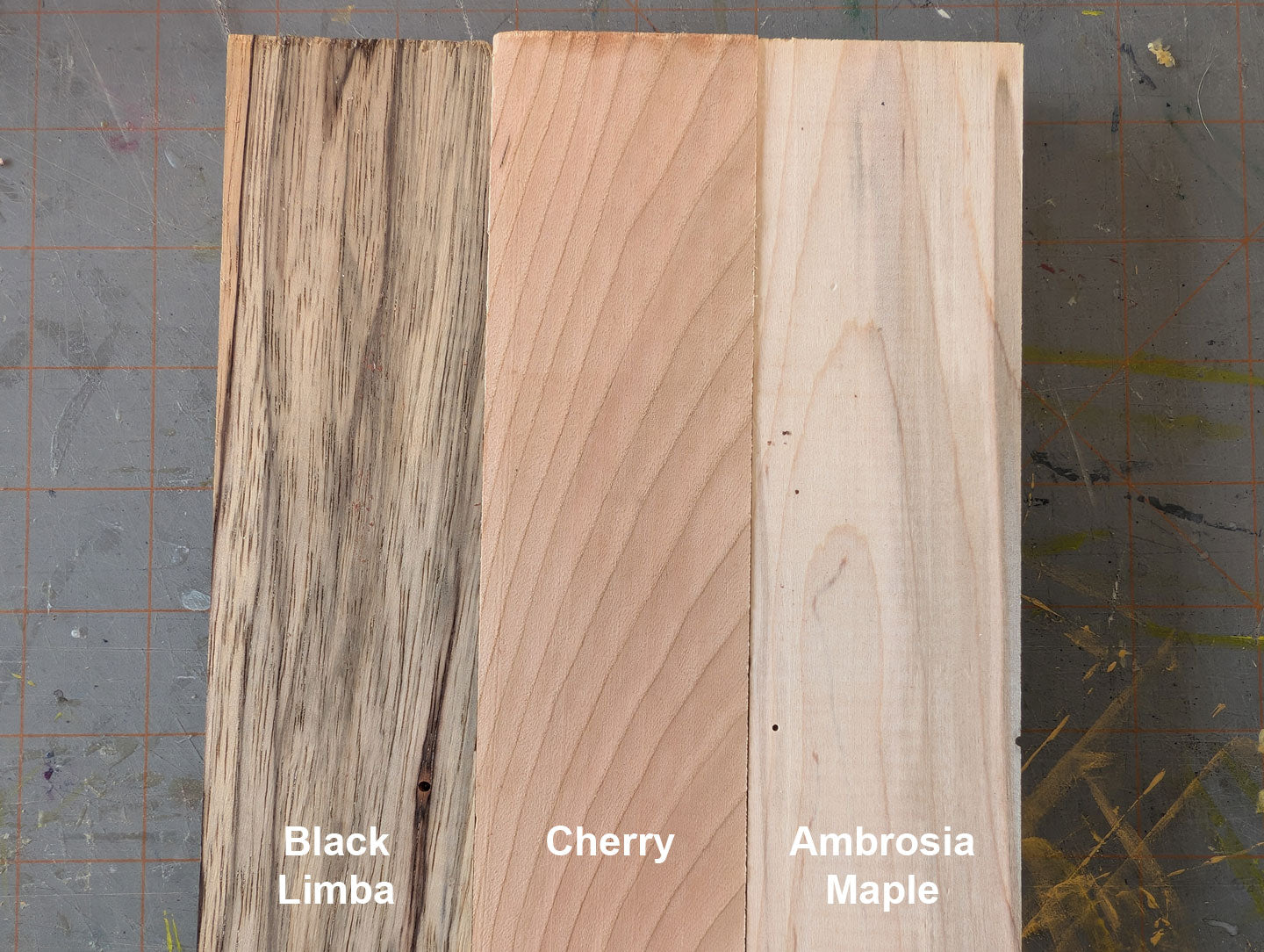 Three wooden planks labeled Black Limba, Cherry, and Ambrosia Maple on a textured surface.